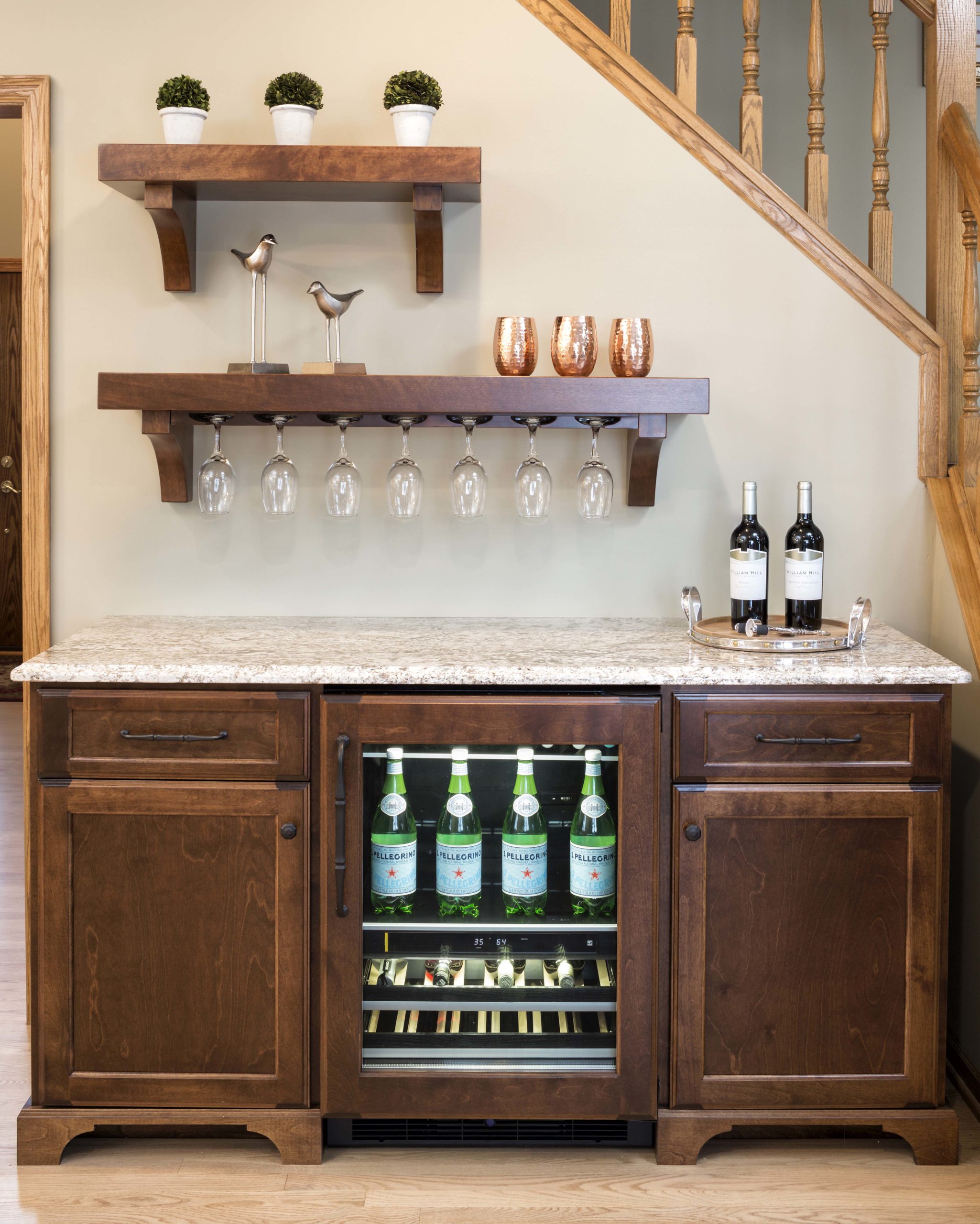 Wine bar with cupboards, small refrigerator, marble countertop and wooden shelf on wall with hanging long stem glasses.