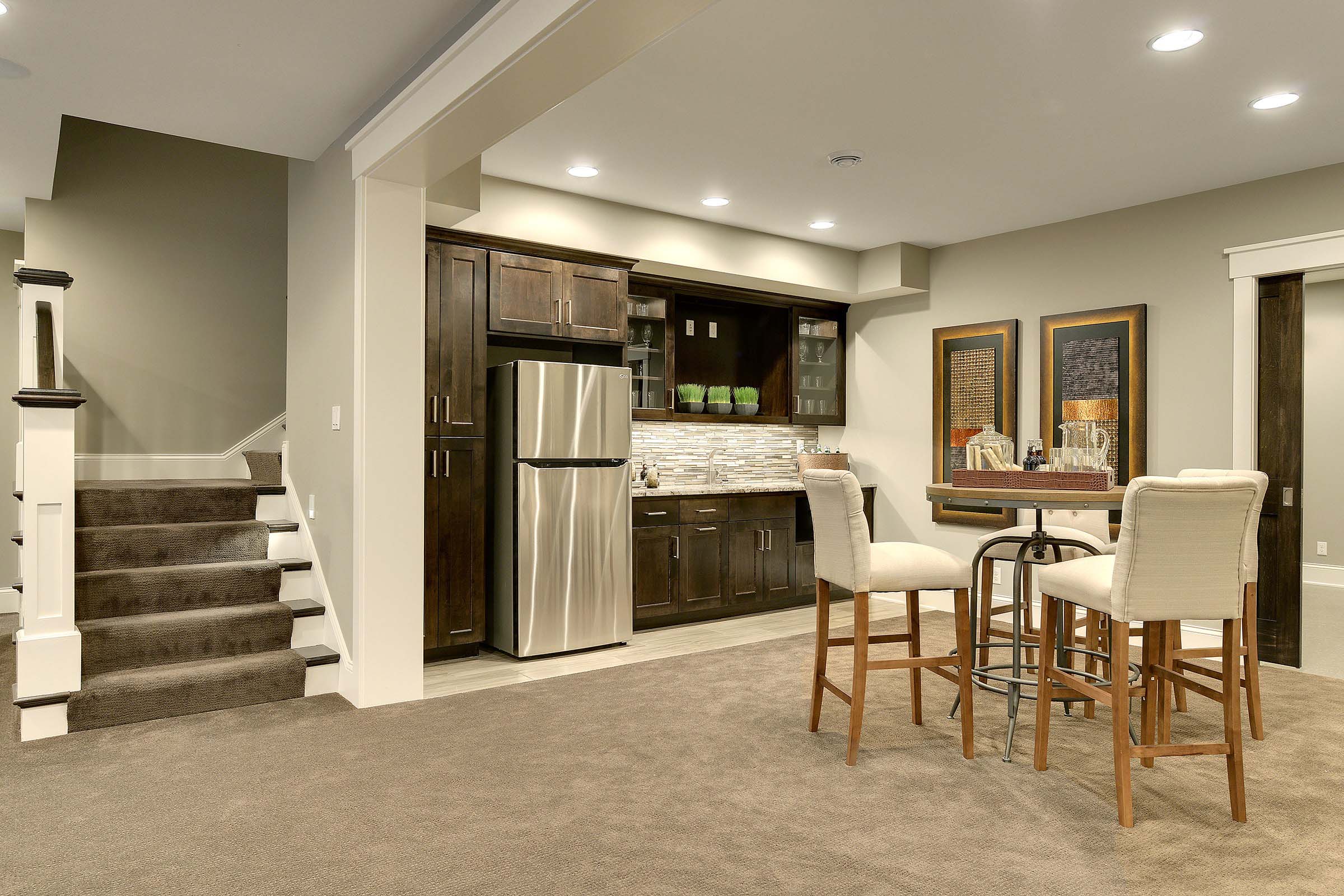 Modern lower level with kitchen area, hightop table seating and carpeted stairs to upper level.