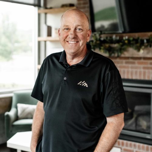 A man in a black polo shirt standing in front of a fireplace.