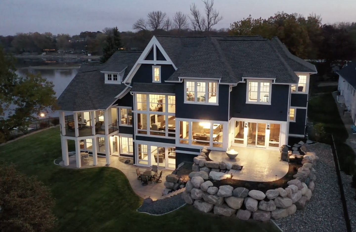 An aerial view of a spacious two-story Prior Lake estates lake home with illuminated interiors and a backyard facing the serene waters at dusk.