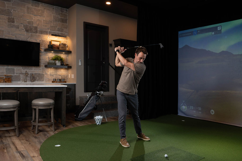 A man practicing golf indoors with a golf simulator and bar remodel in the background.