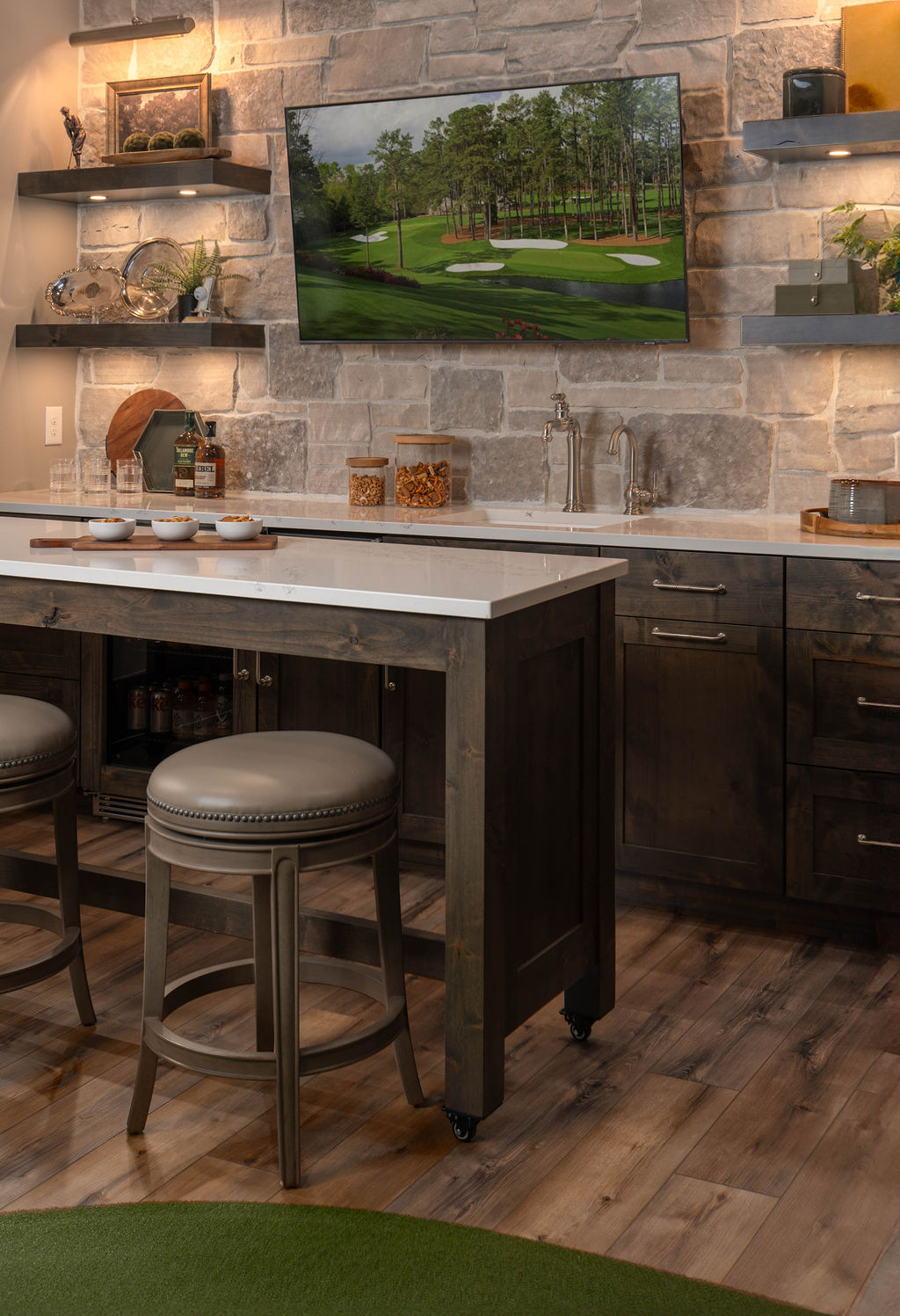 A modern kitchen interior with stone walls, wooden cabinetry, bar stools, and a golf simulator.