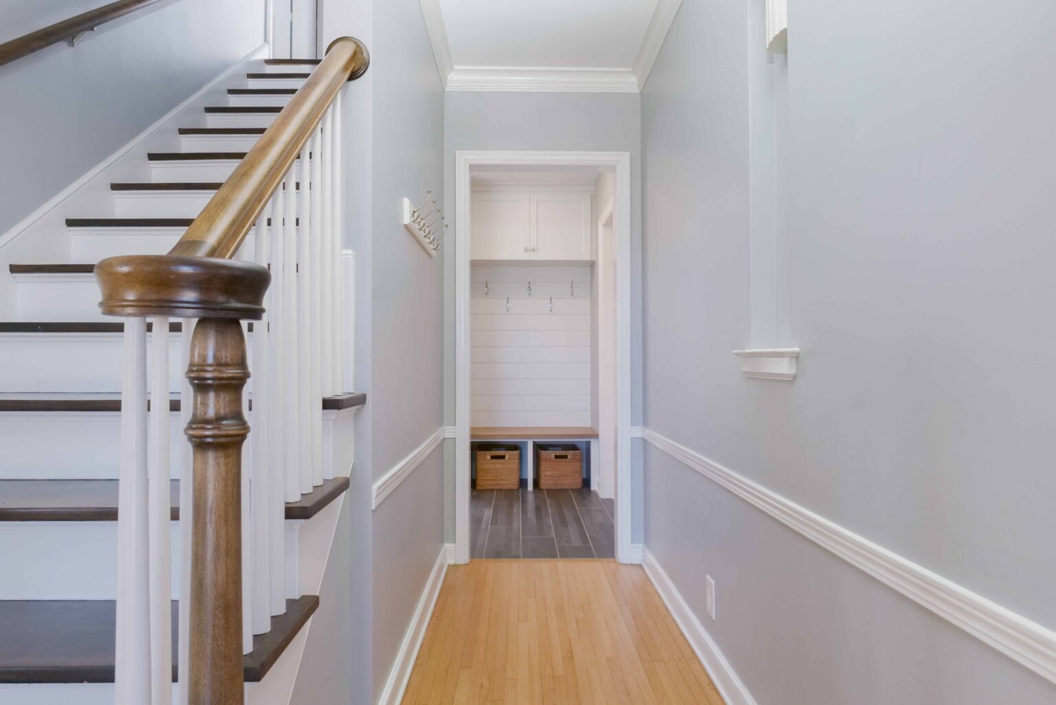 A hallway with wooden flooring leads to a room with hooks and baskets. A staircase with a wooden banister is on the left. Walls are light grey with white trim, showcasing the elegance of the Hampshire Drive remodel.