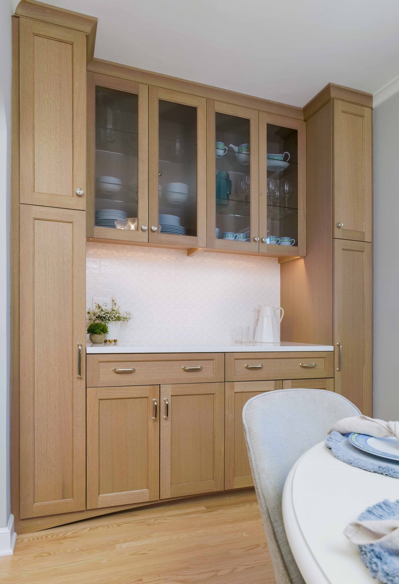 A Hampshire Drive remodel showcases a kitchen cabinet with wooden doors and glass upper panels displaying dishes. Below, the cabinet features a white countertop, drawers, and cabinets with silver handles. A small plant and white jug adorn the counter.