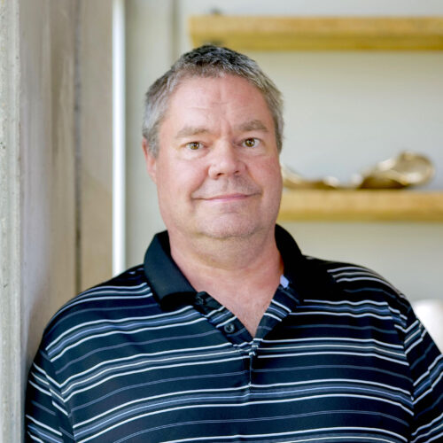 A man with short gray hair and a black striped polo shirt stands indoors, leaning against a wall. Shelves with objects are visible in the background, perhaps reflecting his connection to craftsmanship or details about Highmark Builders.