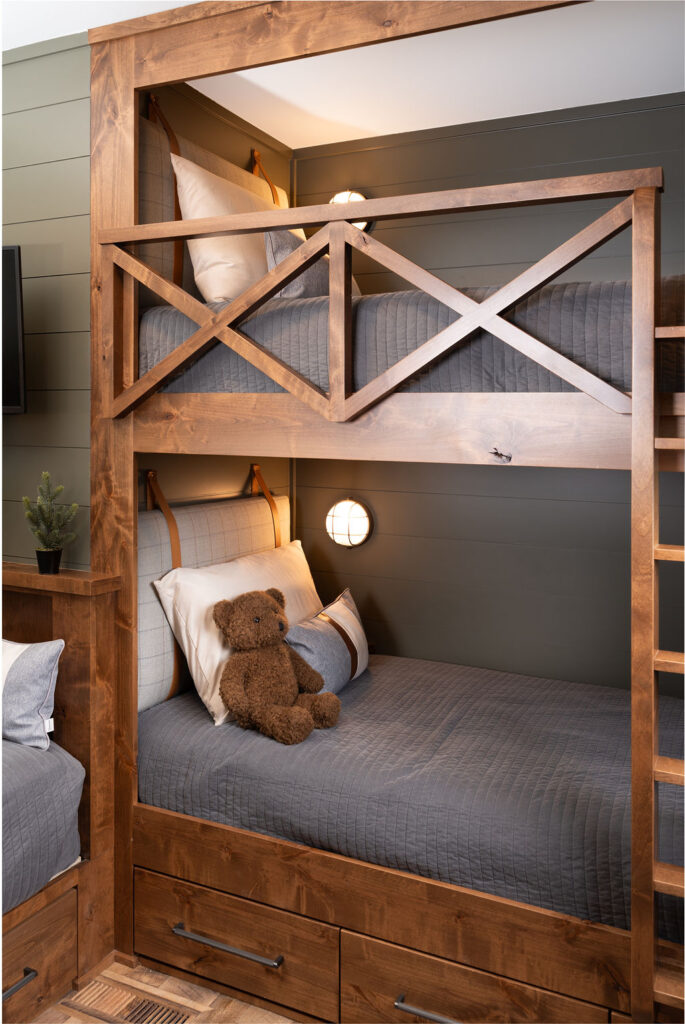 A wooden bunk bed with gray bedding, a teddy bear on the lower bunk, and built-in wall lights on the headboards adds a cozy touch to your Balsam Lake Cabin remodel.