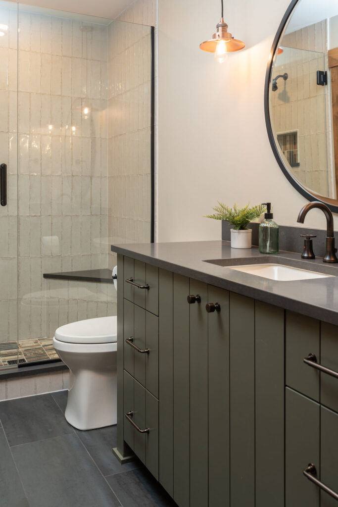 A Balsam Lake Cabin remodel features a modern bathroom with a glass-enclosed shower, white toilet, dark countertop, round mirror, and green cabinets under a single sink.