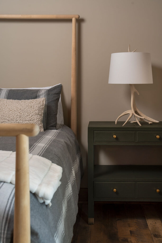 A neatly made bed with striped grey and white bedding sits next to a dark green nightstand with a branch-like lamp on top. The bed features a light wooden headboard, and the room, reminiscent of the Balsam Lake Cabin remodel, has wooden flooring.