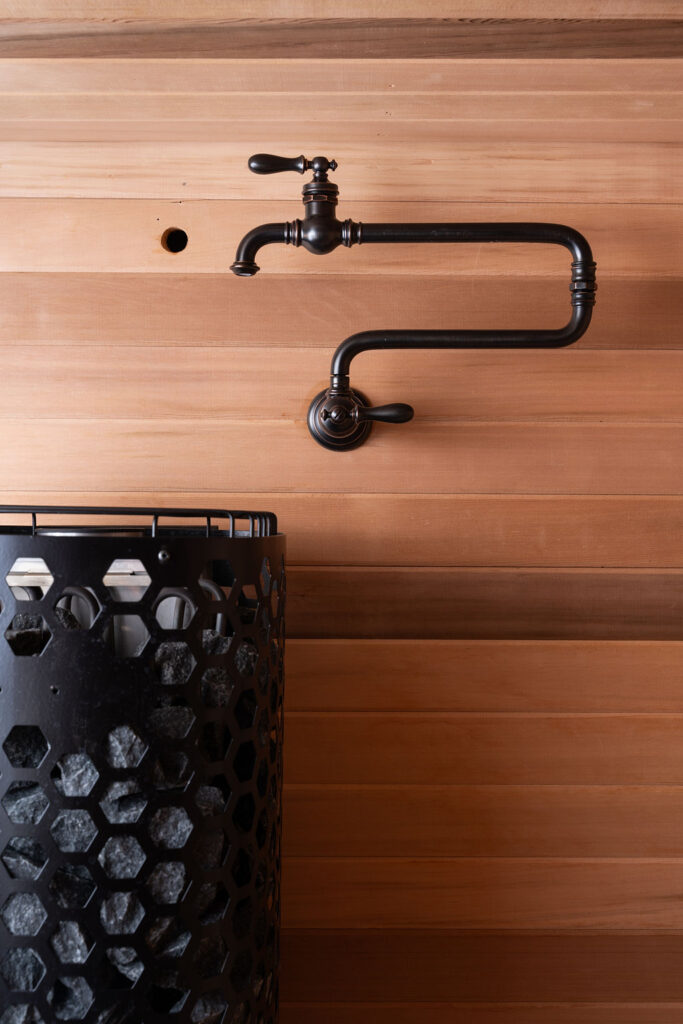 A black sauna heater with hexagonal cutouts stands adjacent to a wall-mounted black faucet against a wooden paneled wall, adding a modern touch to the rustic charm of the Balsam Lake Cabin remodel.