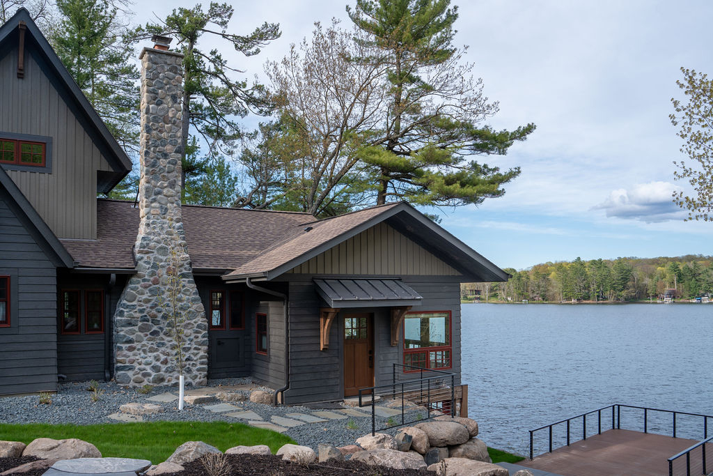 A beautifully remodeled Balsam Lake cabin, featuring a lakeside wooden house with a stone chimney, surrounded by trees and a rocky landscape. A wooden dock extends into the lake in the foreground.