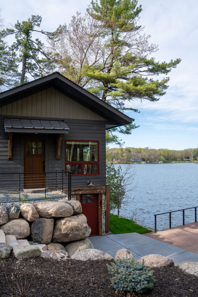 Nestled by Balsam Lake, this remodeled two-story cabin features dark wood paneling and a stone base, accented by a bold red door. Surrounded by trees, the scene is completed with a stone-paved pathway and elegant wrought iron railing.