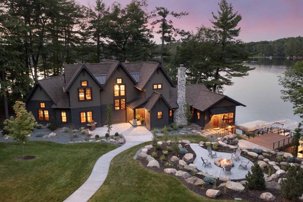 A large dark-colored house, recently part of a Balsam Lake Cabin remodel, is set on the edge of a lake with trees around. The outdoor area features a stone fire pit with seating, a well-maintained lawn, and a pathway.