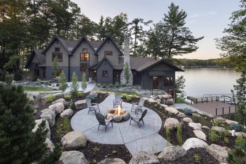 A Balsam Lake cabin remodel includes a lakeside house with a stone fire pit surrounded by chairs, large rocks, and trees in the foreground. A dock extends into the lake, and the house features multiple gabled roofs and large windows.