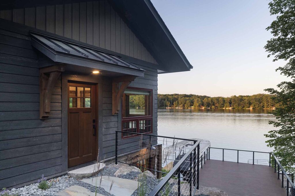 The exterior of the Balsam Lake cabin remodel features a wooden door and deck overlooking a calm lake, surrounded by trees under a clear sky.