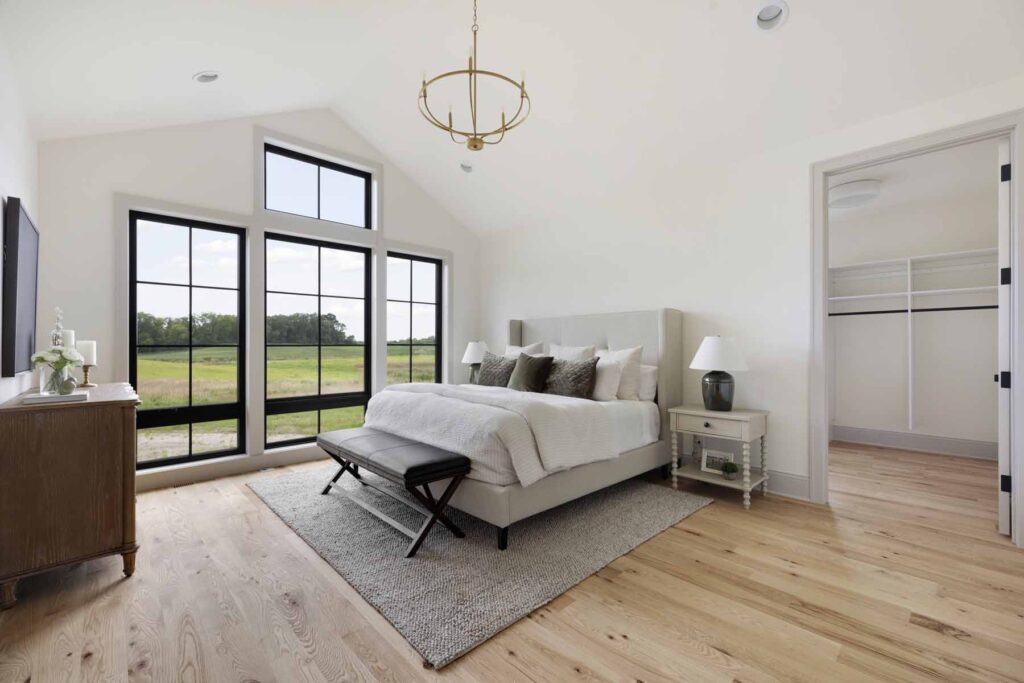 A modern bedroom exuding elegance, featuring a large bed with a bench and side tables on a rug. Large windows offer lush greenery views, while an open closet door adds convenience. Perfect for those exploring properties and homes for sale, the chandelier completes this inviting space.