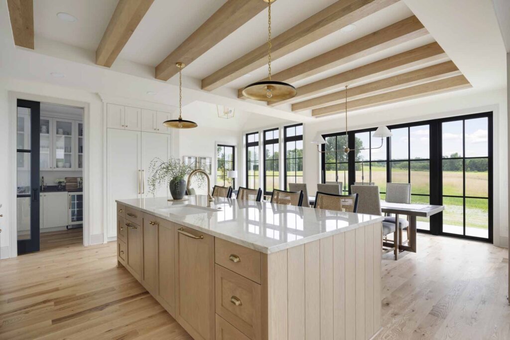A modern kitchen in The Oxbow Ranch Home boasts a large island, wooden beams, pendant lights, and a dining area with floor-to-ceiling windows overlooking a serene field.