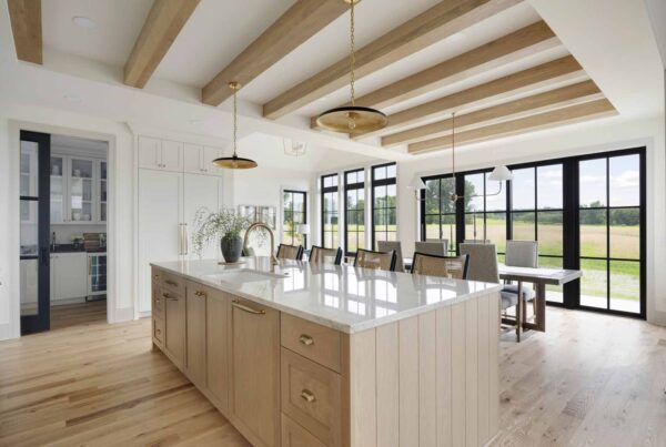 A modern kitchen in The Oxbow Ranch Home boasts a large island, wooden beams, pendant lights, and a dining area with floor-to-ceiling windows overlooking a serene field.