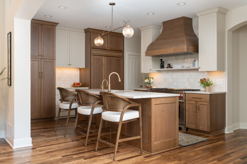 Modern kitchen with wood cabinetry, an island with three chairs, pendant lights, and a range hood. Perfect for those looking to remodel your home, it features plants and fruit on the counters, with hardwood flooring throughout.