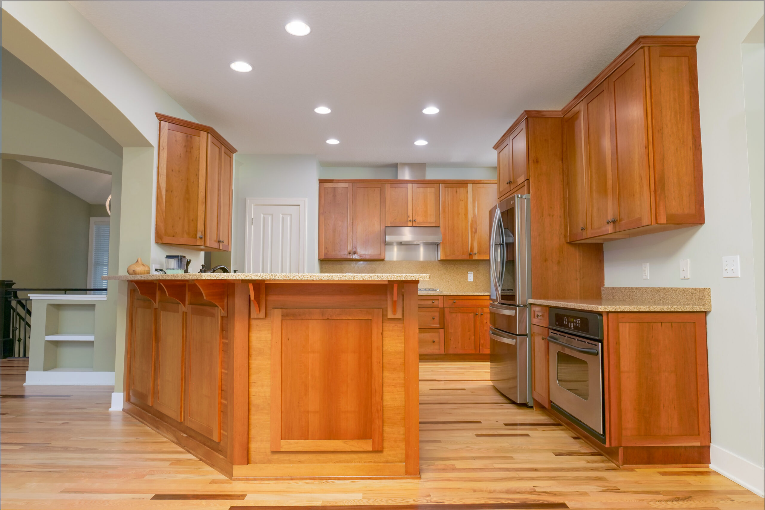 A modern kitchen invites you to remodel your home with its wooden cabinets, stainless steel appliances, and recessed lighting. The room boasts a spacious island and elegant hardwood floors.