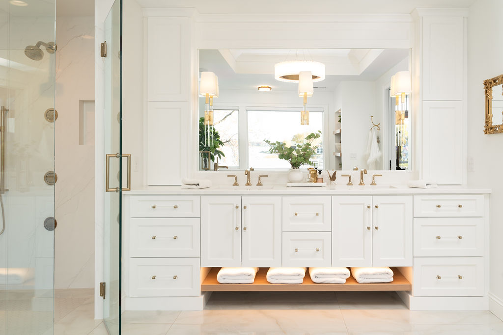 Excelsior bathroom remodel: A pristine white bathroom featuring a double sink with elegant gold fixtures and a large mirror. Below, shelves neatly hold folded towels, while a sleek glass shower door is visible on the left.