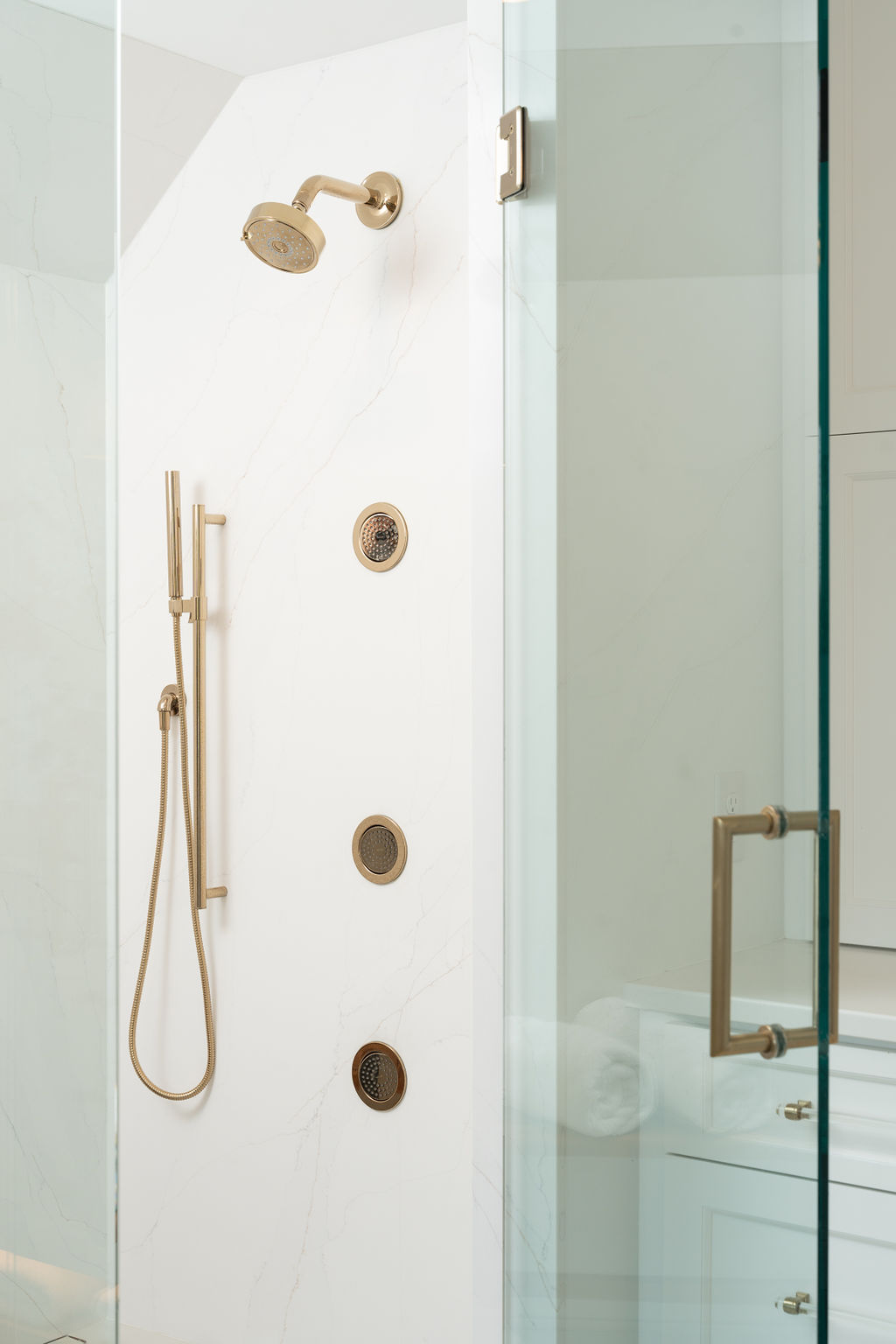 Experience the Excelsior bathroom remodel with a modern shower featuring gold fixtures—a stylish showerhead, versatile handheld spray, and three invigorating wall jets—all elegantly set against a white marble backdrop and enclosed by sleek glass.