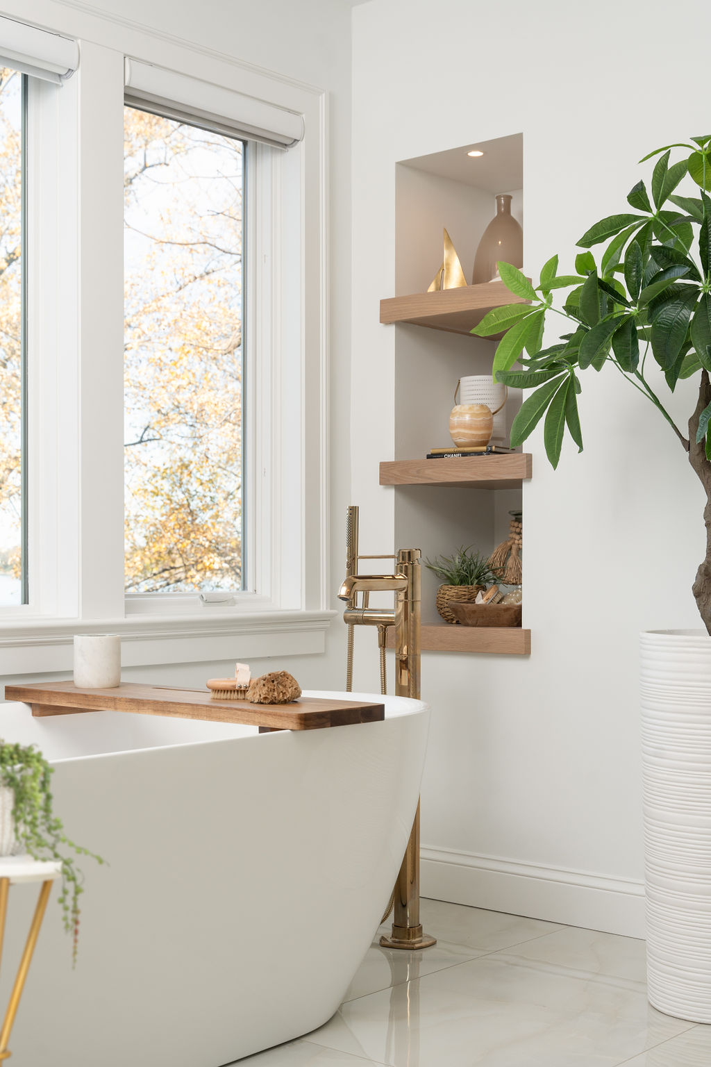 Excelsior bathroom remodel featuring a modern design with a freestanding tub, large window, and wooden shelves displaying decor items. A plant rests near the tub, complemented by a gold faucet for an elegant touch.