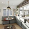 A bright south Minneapolis kitchen remodel features large windows, green cabinets, and a farmhouse sink. In the corner, a small dining area with a table and bench seating adds charm. Plants and kitchenware adorn the inviting space.